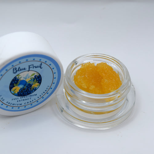A small clear glass jar filled with bright yellow-orange crystalline powder sits next to its white lid, which features a blue and yellow 'Blue Fruit' label, on a plain white background.