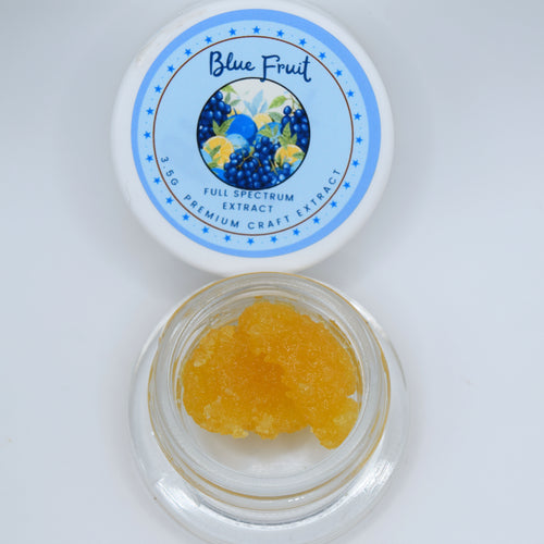 An open clear jar filled with yellow-orange crystalline powder sits below its white lid, which features a blue circular label reading 'Blue Fruit Full Spectrum Extract'.