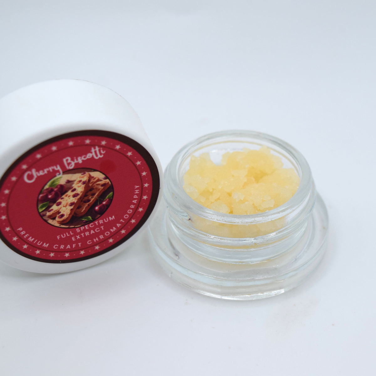 An open clear glass jar filled with pale yellow crystalline product next to its white lid, which features a red circular label reading 'Cherry Biscotti' and 'Full Spectrum Premium Craft Cholesterol & Spermidin'.