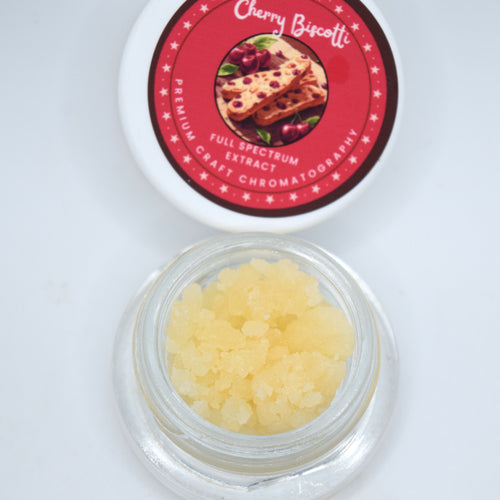 An open clear jar filled with pale yellow crystalline scrub sits below its white lid, which has a red circular label reading Cherry Biscotti.