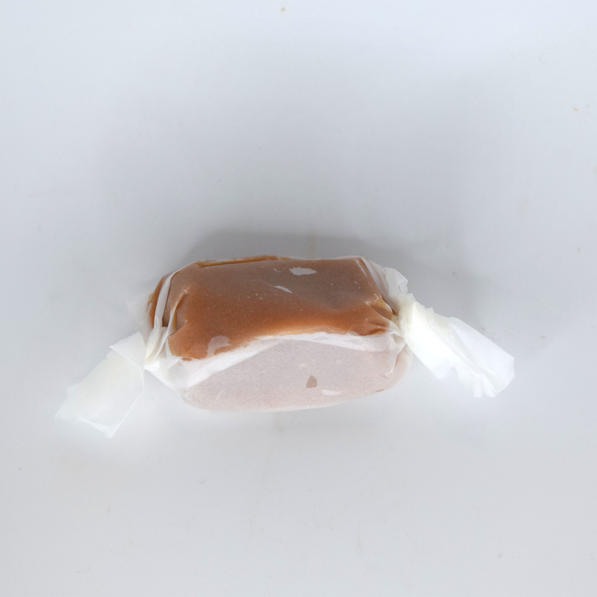 A single wrapped candy or caramelly piece on a plain white background, with a glossy brown top and pale pinkish-beige sides wrapped in white paper. Soft 50mg Nano Delta-9 infused caramel made with sugar and evaporated milk for eating directly or dissolving into coffee.