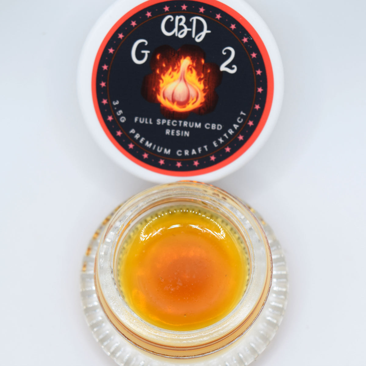 A small clear glass jar filled with amber-orange resin sits below its white lid, which features a black circular label with red stars and a flame graphic. The label reads 'CBD G2' and 'FULL SPECTRUM CBD RESIN'.
