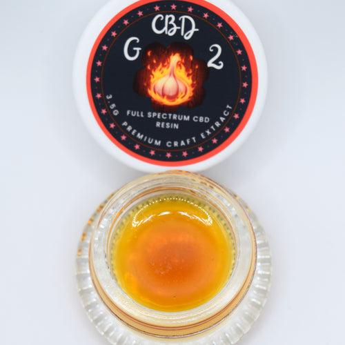 A small clear glass jar filled with amber-orange resin sits below its white lid, which features a black circular label with red stars and a flame graphic. The label reads 'CBD G2' and 'FULL SPECTRUM CBD RESIN'.