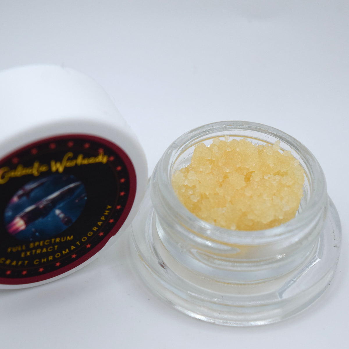 A small clear glass jar filled with pale yellow crystalline powder or extract, shown next to its white lid with a dark circular label reading Galactic Warheads.