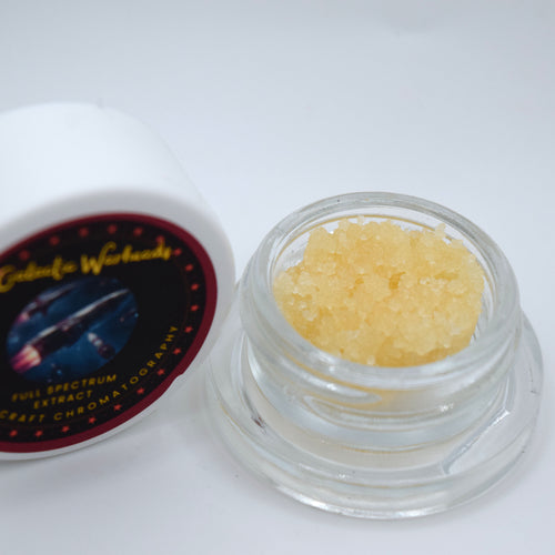 A small clear glass jar filled with pale yellow crystalline powder or extract, shown next to its white lid with a dark circular label reading Galactic Warheads.