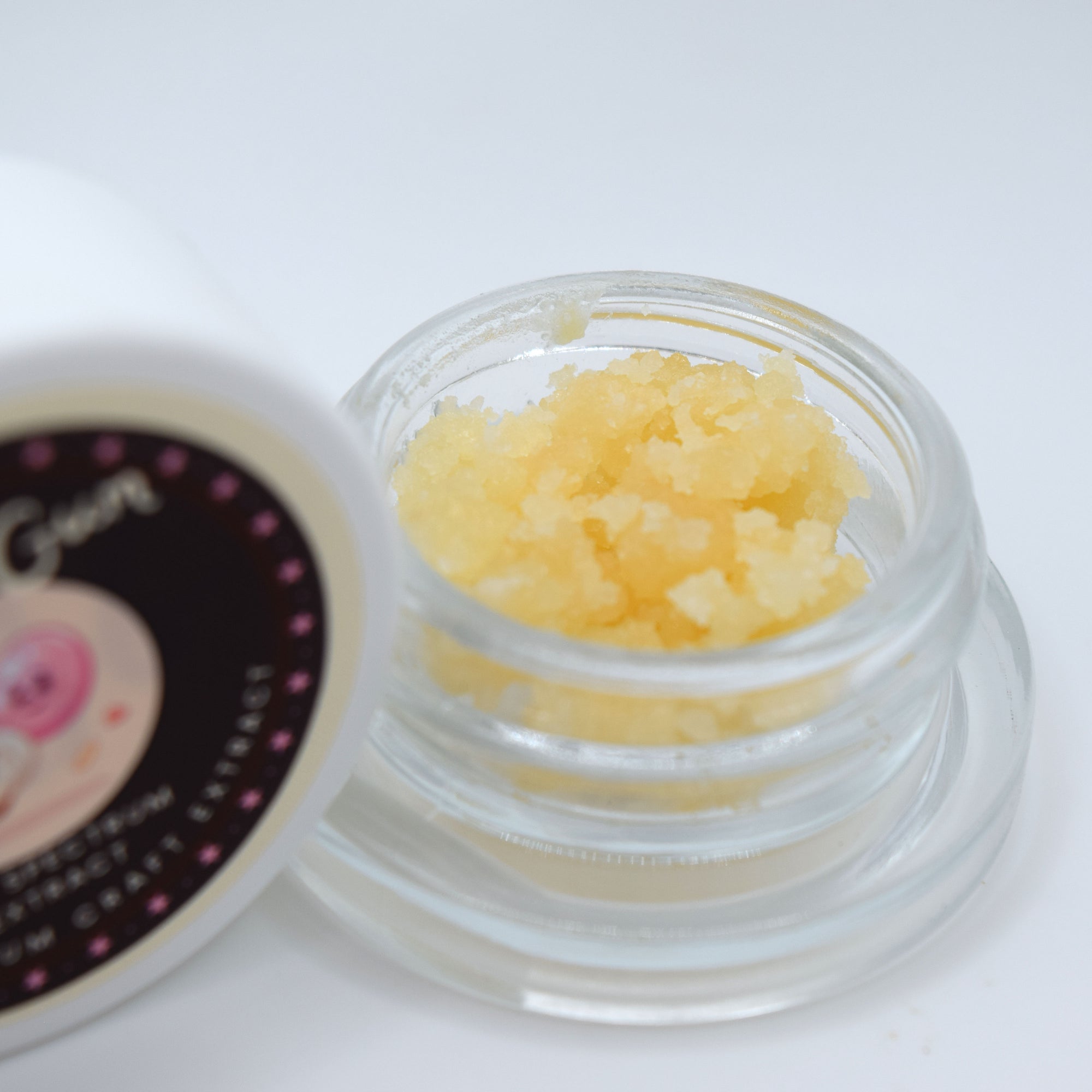 A small clear glass jar filled with pale yellow granular scrub, with the white lid partially visible and a decorative dark label on the left, photographed on a clean white background.