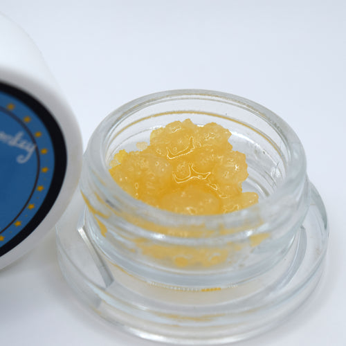 A close-up of a small clear glass jar filled with yellow translucent caviar, with part of a white lid featuring a blue label visible on the left against a white background.