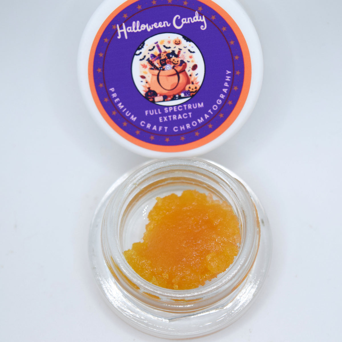 An open clear glass jar containing orange crystalline powder or granules, with the white lid placed above it. The lid has a purple circular label reading 'Halloween Candy' and 'Full Spectrum Extract' with a pumpkin illustration.