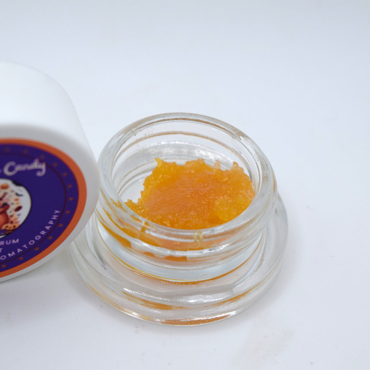 An open clear glass jar containing a textured orange gel or wax, with part of a white container lid visible on the left. The lid has a purple and orange label with partial text including 'Candy' and 'MUMATOMATOGRAPH'.