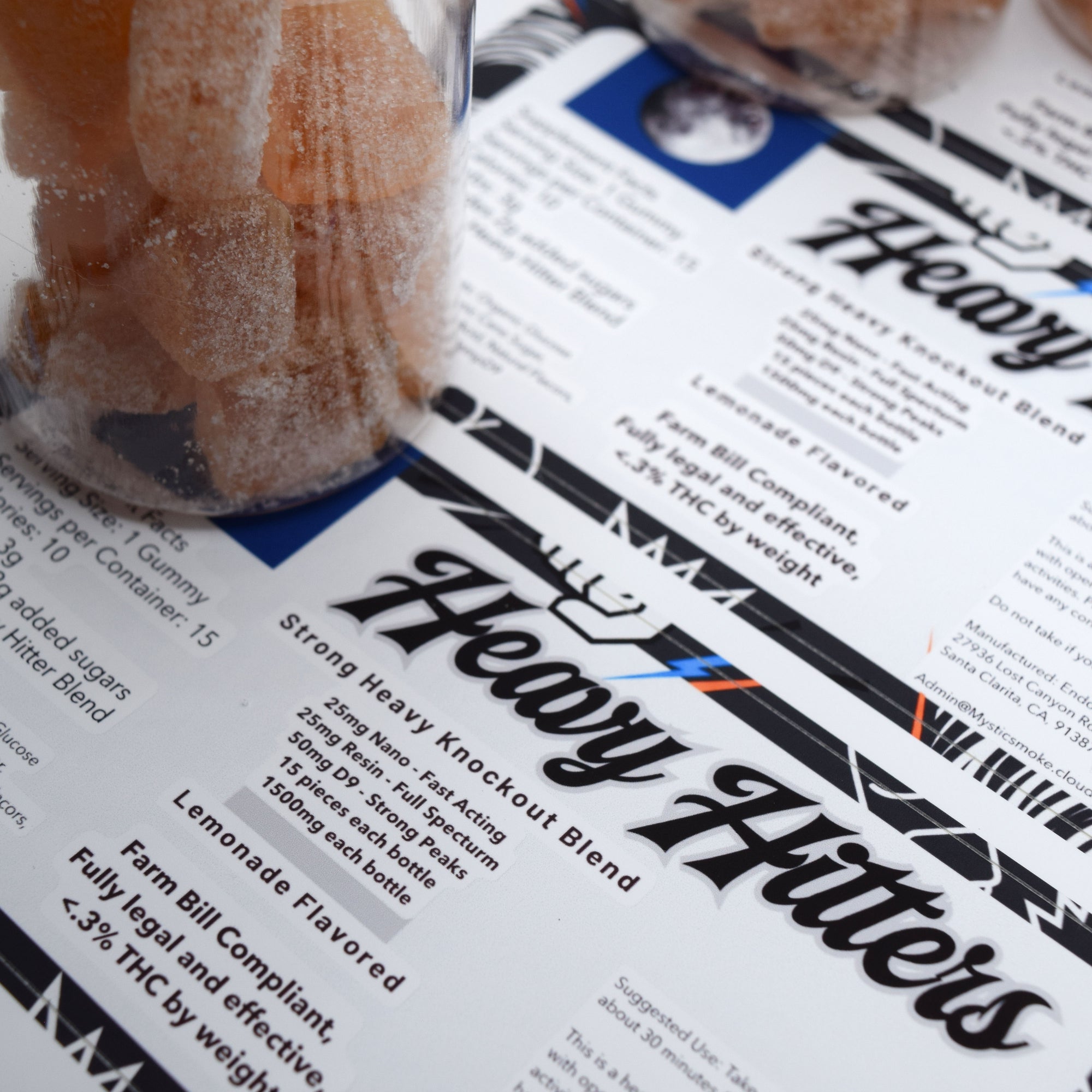 A close-up of printed product information cards for a cannabis strain called Heavy Hitters, with a glass jar of gummy candies partially visible on a white surface.