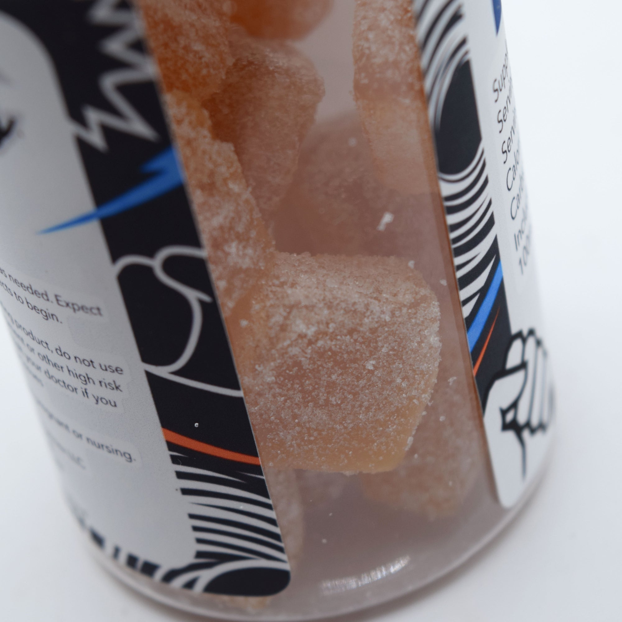 A close-up of a clear plastic jar filled with translucent orange-brown gummy candies coated in white sugar, with a black, white, and blue label partially visible.