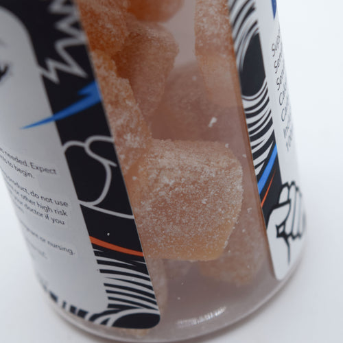 A close-up of a clear plastic jar filled with translucent orange-brown gummy candies coated in white sugar, with a black, white, and blue label partially visible.