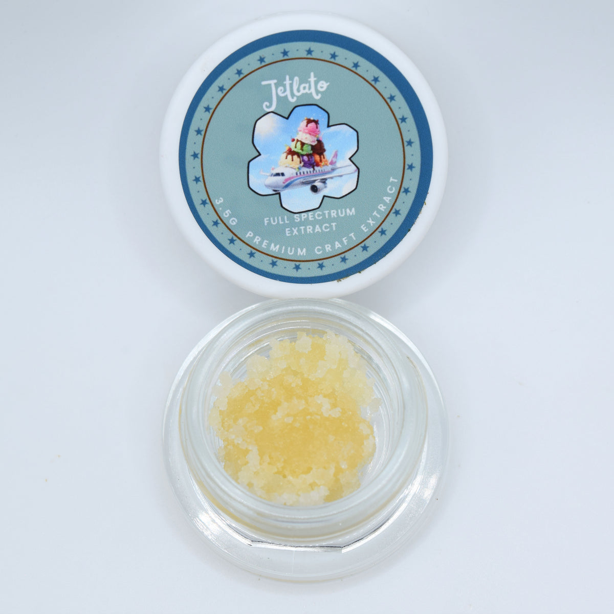 An open round clear plastic jar containing pale yellow crystalline material, shown below its matching white lid which features a blue circular label for 'Jettato Full Spectrum Extract'.