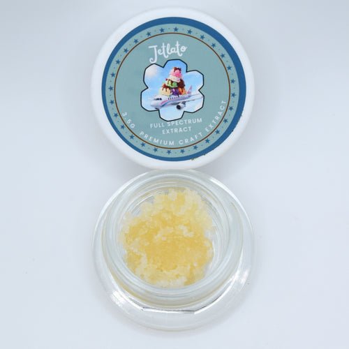 An open round clear plastic jar containing pale yellow crystalline material, shown below its matching white lid which features a blue circular label for 'Jettato Full Spectrum Extract'.