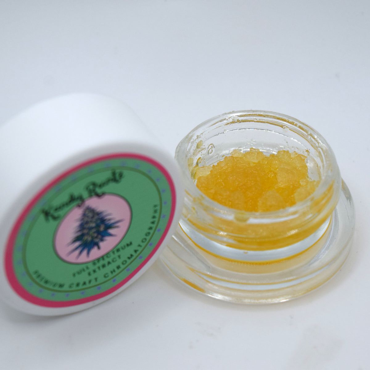 A small clear glass jar filled with yellow-orange crystalline powder, shown next to its white lid which features a green, pink, and blue label for 'Karma Roots' full-spectrum hemp craft chroma gel.