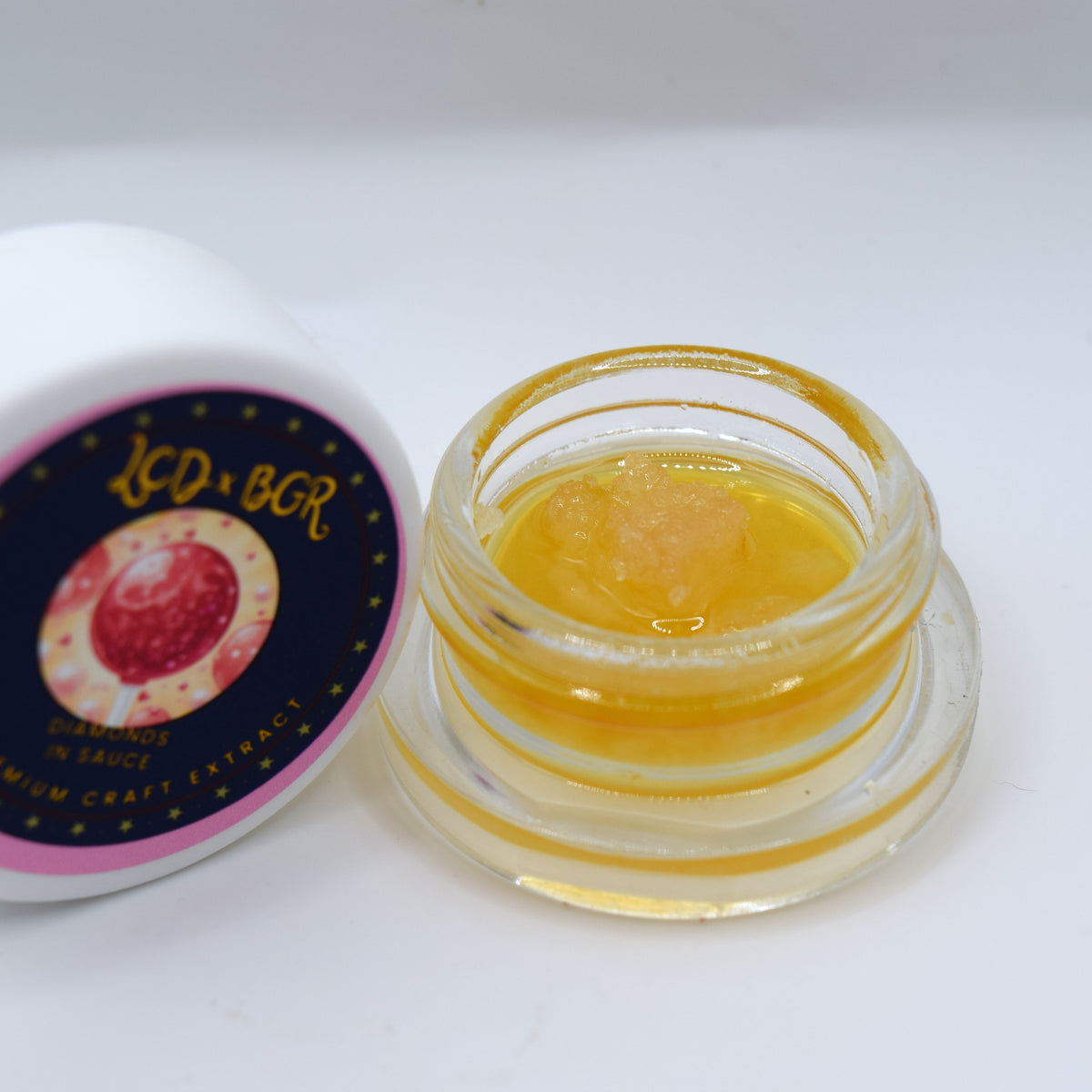 A small clear glass jar filled with golden yellow balm or extract, shown next to its white lid which features a dark navy label with gold lettering and a pink circular graphic.