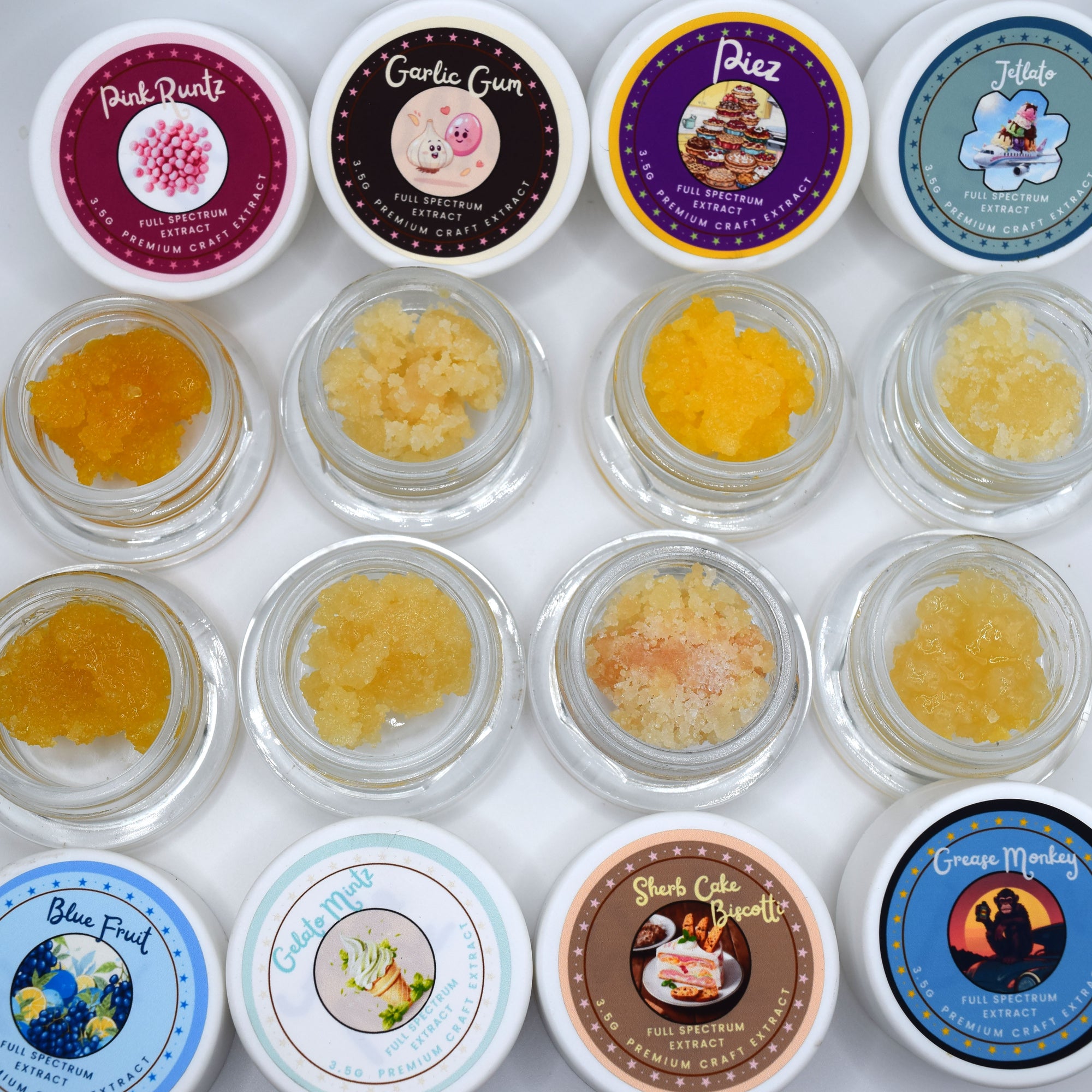 Twelve small clear jars filled with textured, granular bath salts are arranged in a grid with their colorful lids visible above each. The lids feature various illustrated labels with names including Pink Pearl, Garlic Gun, Piez, Tjate, Blue Fruit, Cinnamon Apple, Yellow Sun, and Goose Meadow.