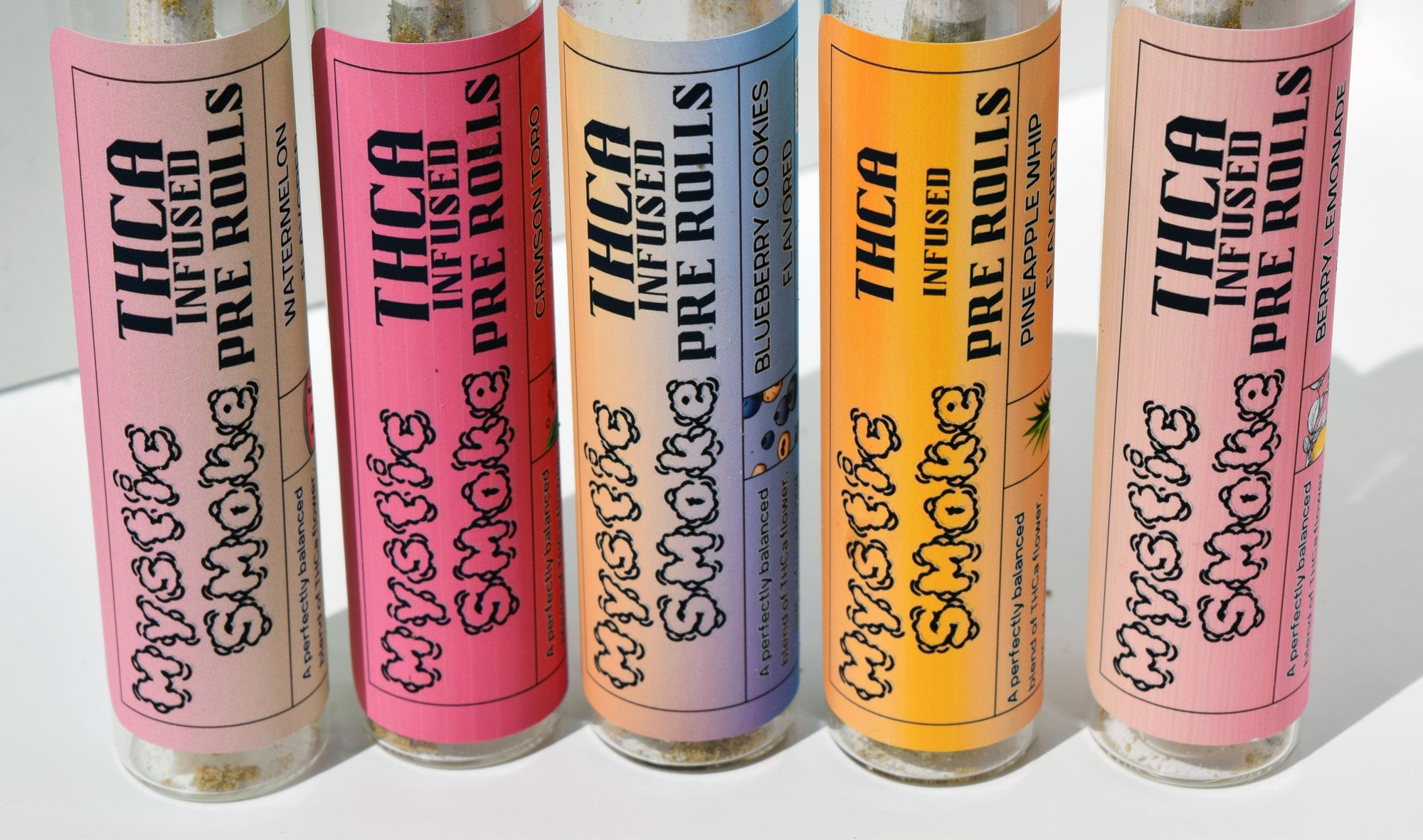 Five clear glass tubes of pre rolls with colorful labels in pink, red, blue, orange, and light pink, arranged in a row on a white surface. The labels read 'Mystic Smoke' and 'THCA Infused / Flavored Pre Rolls' with different flavor names including Watermelon, Blueberry Cookies, Pineapple Sand, and Blueberry Jam.