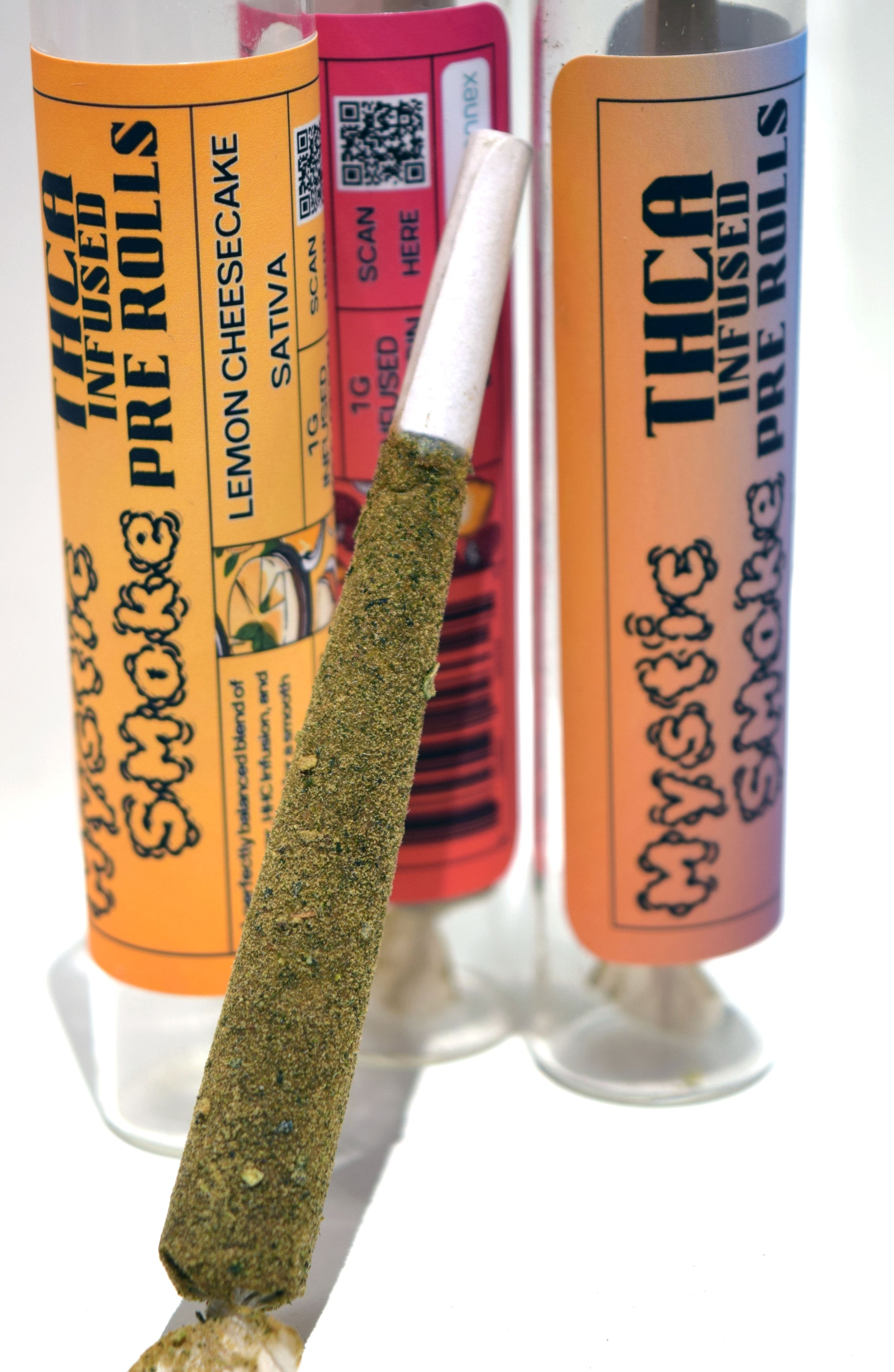 A close-up product shot of pre-rolled joints and labeled packaging on a white background. The front shows a rolled joint with coarse green herb, with orange and pink labels reading 'THCA INFUSED PRE ROLLS' and 'Mystic Smoke'.