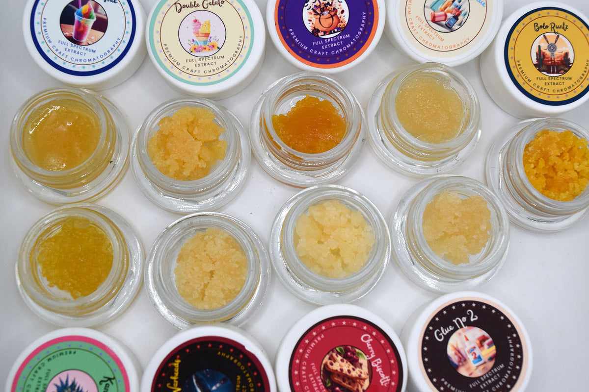 An overhead grid of small clear jars containing textured yellow and orange cosmetic or bath salts, arranged on a white background with colorful round lids visible around the edges.