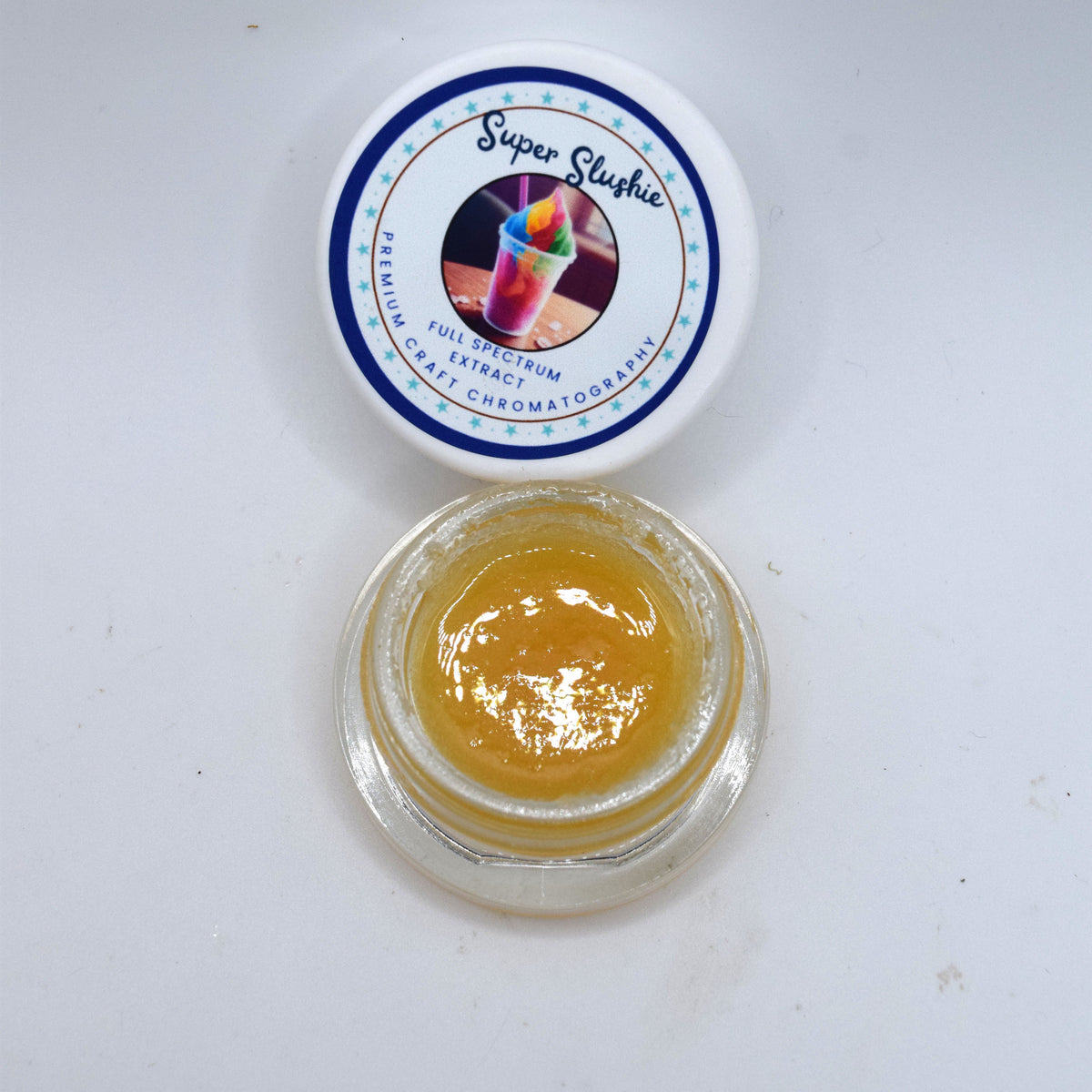 An open clear jar containing a thick golden yellow gel or balm, with its white lid placed above it. The lid has a printed label reading 'Super Slushie' with a colorful drink illustration and blue decorative border.