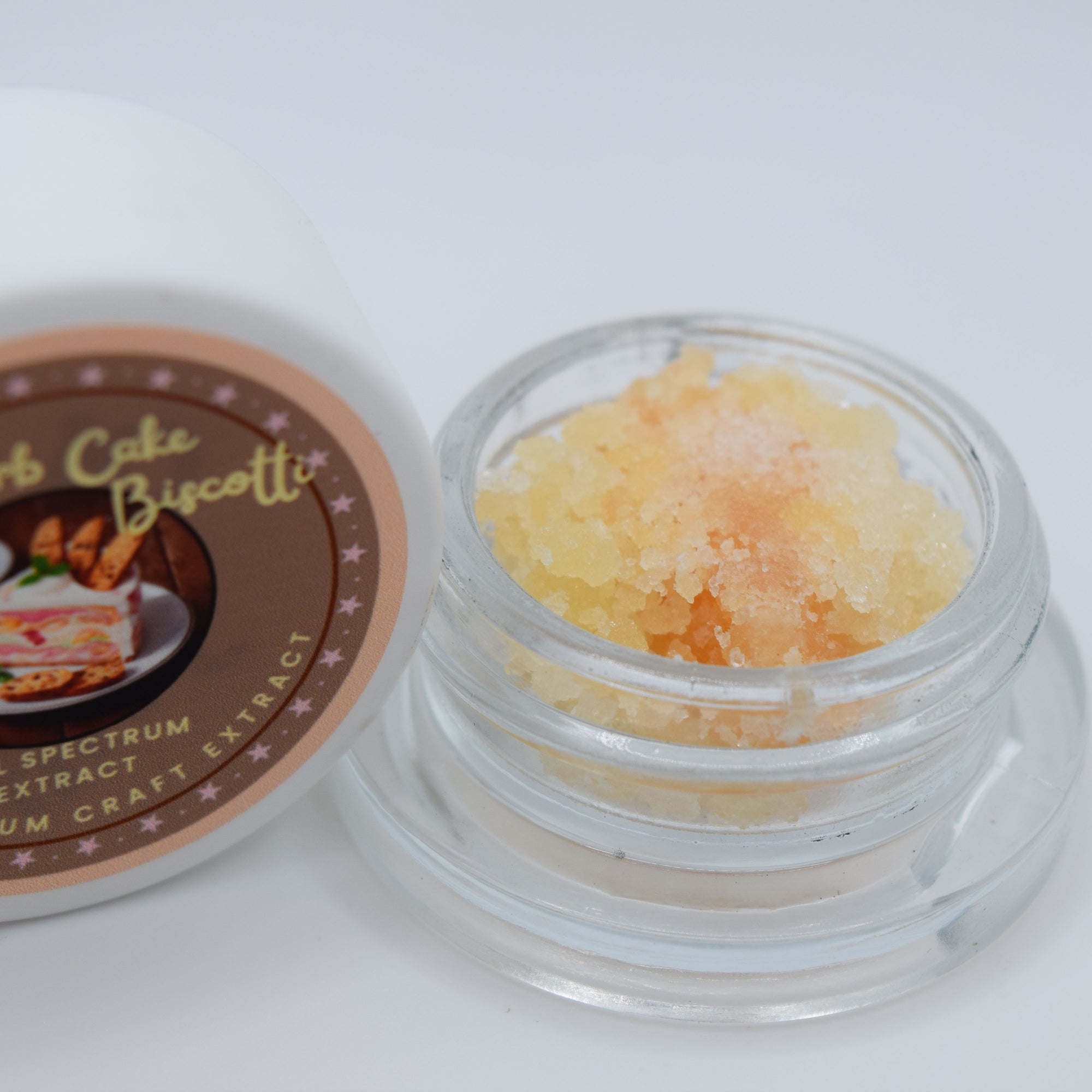 An open clear jar filled with pale yellow-orange crystalline extract next to its white lid, which features a brown label reading 'Creme Cake Biscotti' and 'Spectrum Craft Extract'.