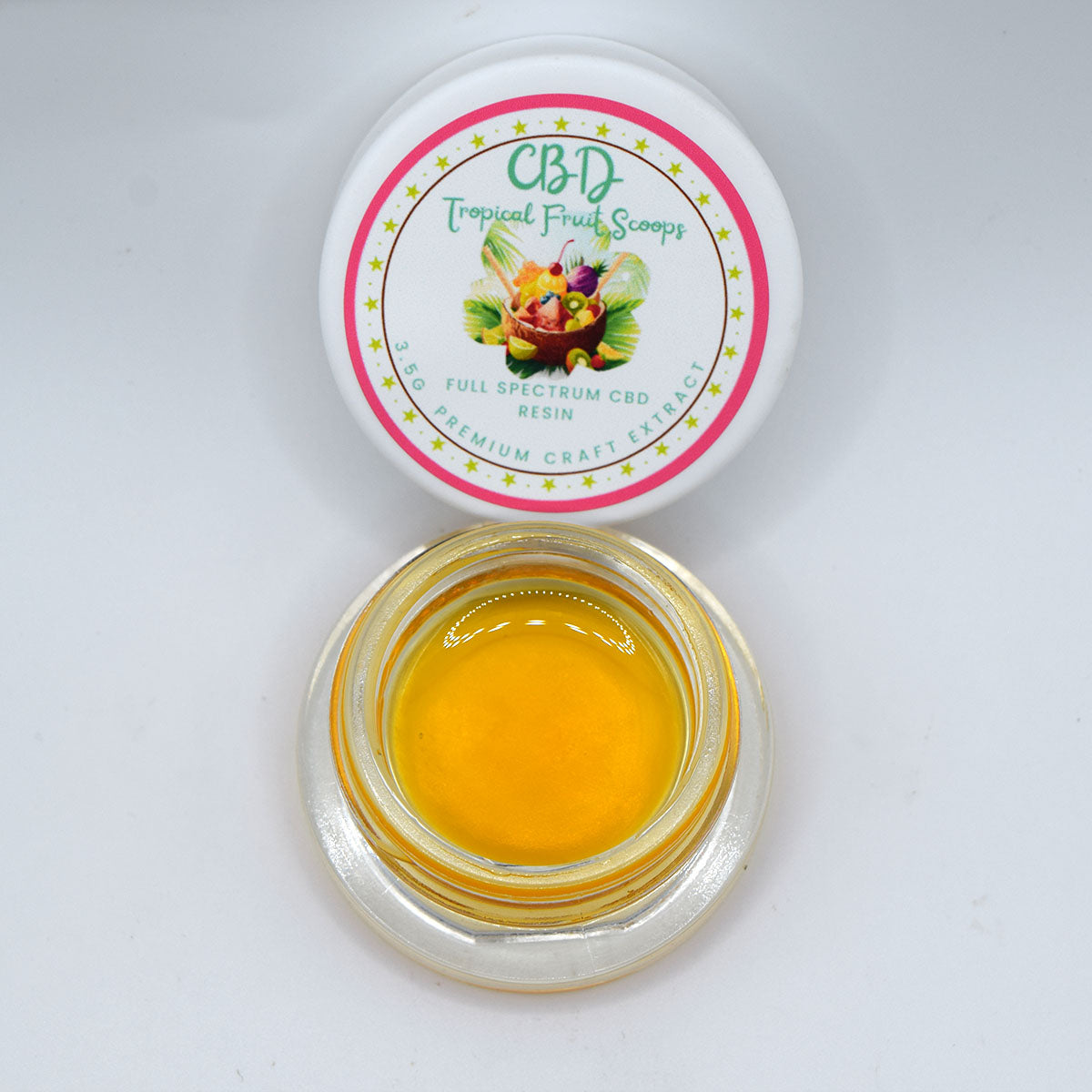 An open clear glass jar filled with bright yellow-orange CBD resin, shown below its white lid which features a colorful tropical fruit label reading 'CBD Tropical Fruit Scoops'.