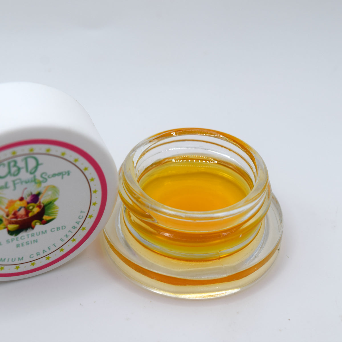 An open clear glass jar filled with golden amber resin, shown next to its white lid which features a colorful label for 'CBD Gummy Scops' on a plain white background.